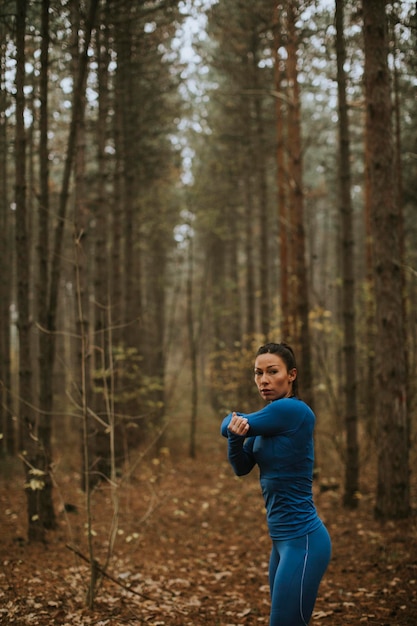 Hübsche junge Frau im blauen Trainingsanzug, die sich vor dem Training im Herbstwald ausdehnt