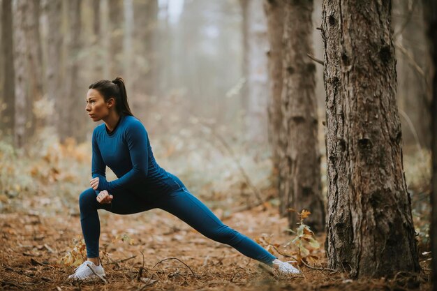 Hübsche junge Frau im blauen Trainingsanzug, die sich vor dem Training im Herbstwald ausdehnt