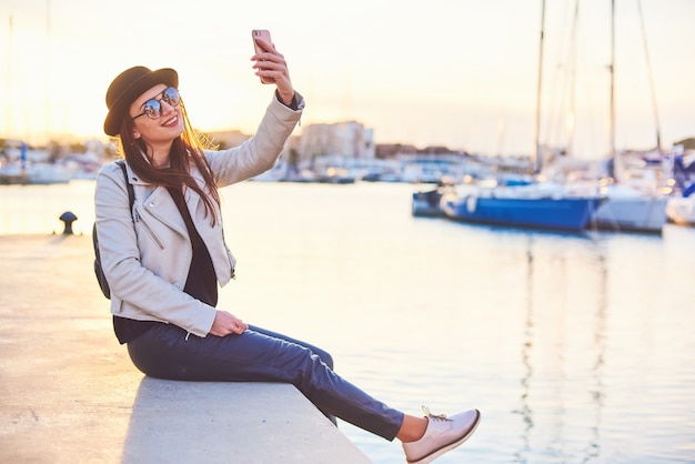 Hübsche junge Frau im schwarzen Hut, die auf einem Pier mit einem Telefon an einem Seehafen sitzt