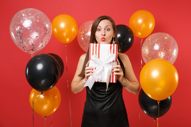 Hübsche junge Frau im schwarzen Kleid, die feiert, rote Schachtel mit Geschenk hält, bläst Lippen präsent, Luftkuss auf rotem Hintergrund Luftballons sendend. Frohes neues Jahr, Geburtstagsmodell Urlaub Partykonzept.