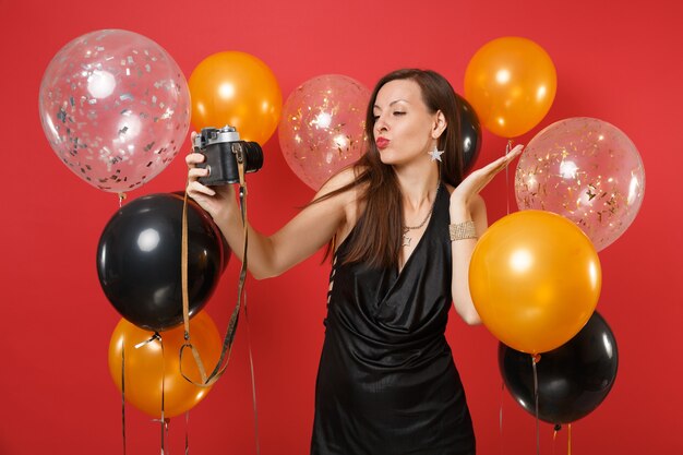 Hübsche junge Frau im schwarzen Kleid macht Selfie-Aufnahme auf Retro-Vintage-Fotokamera, bläst Lippenküsse auf leuchtend roten Luftballons. Happy New Year Geburtstag Mockup Urlaub Party Konzept.