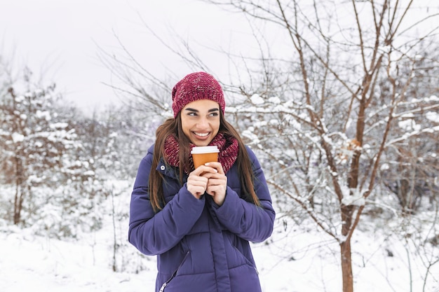 Hübsche junge Frau im Winteroutfit, die Einwegbecher mit heißem Kaffee oder Tee gefüllt hält.