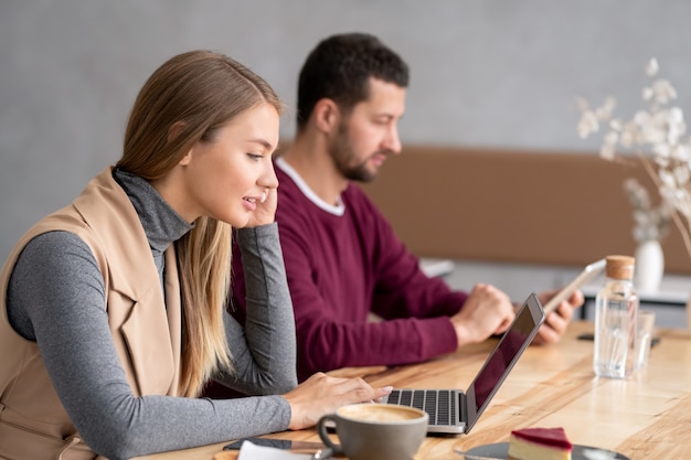 Hübsche junge Frau in der Freizeitkleidung, die durch Holztisch vor Laptop-Anzeige sitzt und durch Video-Chat kommuniziert