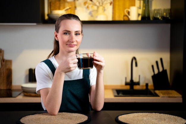 Hübsche junge Frau in einer grünen Küchenschürze mit einer Tasse Kaffee in den Händen sitzt im Kitc