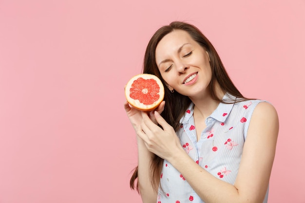 Hübsche junge Frau in Sommerkleidung, die die Augen geschlossen hält und die Hälfte der frischen, reifen Grapefruit isoliert auf rosa Pastellhintergrund hält. Menschen lebendiger Lebensstil entspannen Urlaubskonzept. Mock-up-Kopienbereich.