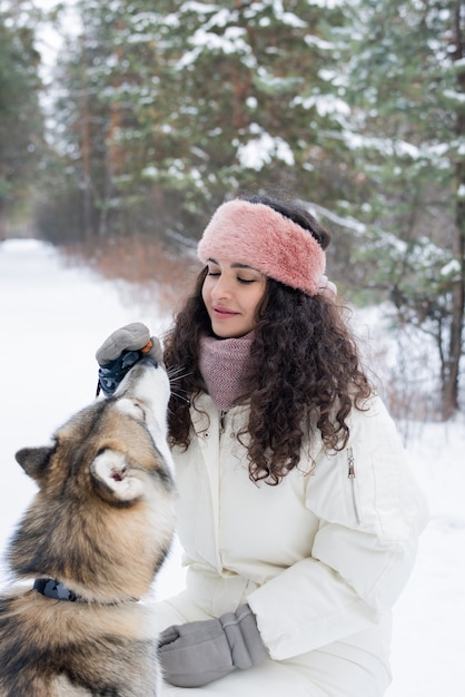 Hübsche junge Frau mit dunklem langem lockigem Haar, das reinrassiges siberianisches husky Haustier füttert, während es im verschneiten Park kühlt