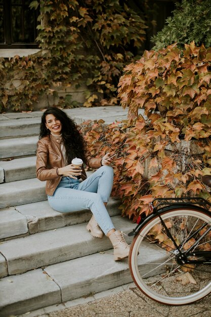 Hübsche junge Frau mit Handy trinken Kaffee zum Mitnehmen an der Treppe mit dem Fahrrad am Herbsttag