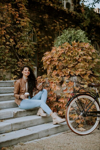 Hübsche junge Frau mit Handy trinkt Kaffee, um am Herbsttag mit dem Fahrrad auf die Treppe zu gehen