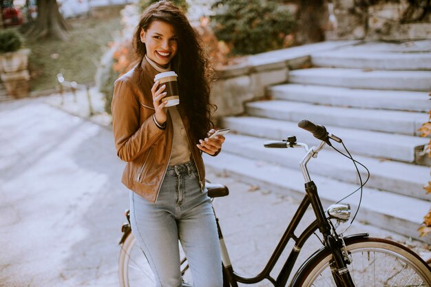 Hübsche junge Frau mit Handy trinkt Kaffee, um am Herbsttag mit dem Fahrrad zu fahren