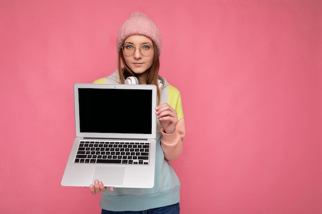 Hübsche junge Frau mit Hut, buntem Pullover und Brille, die einen Computer-Laptop mit leerem Kopienraum-Bildschirm hält, der weiße Kopfhörer trägt und die Kamera einzeln auf rosafarbenem Wandhintergrund betrachtet