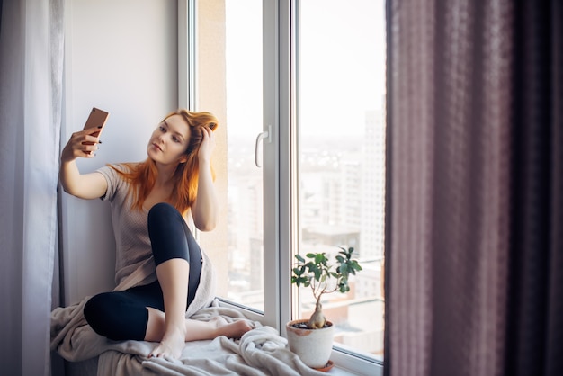 Hübsche junge Frau mit langen roten Haaren macht ein Selfie auf ihrem Smartphone, während sie auf der Fensterbank sitzt. Schönes Mädchen mit einem Telefon in einer häuslichen Umgebung. Konzept für Technologie und soziale Netzwerke.