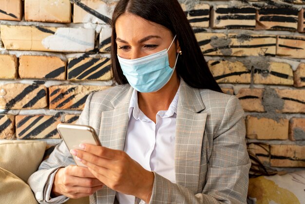 Hübsche junge Frau mit schützender Gesichtsmaske unter Verwendung des Mobiltelefons im Café