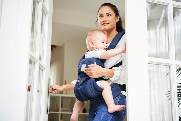 Hübsche junge gemischtrassige Frau, die die Eingangstür öffnet und das Haus mit dem Baby in der Trage verlässt