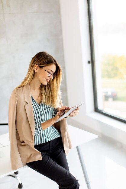 Hübsche junge Geschäftsfrau, die digitales Tablett hält und im modernen Büro steht