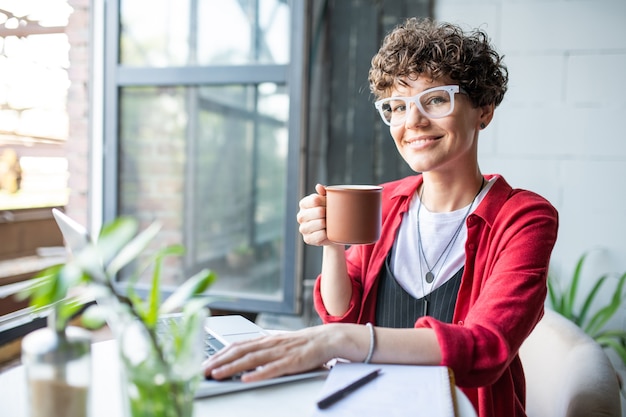 Hübsche junge glückliche Frau in der intelligenten Freizeitkleidung, die Sie beim Tee oder Kaffee betrachtet und Laptop im Café verwendet