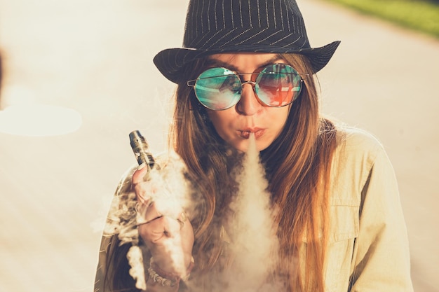 Hübsche junge Hipster-Frau mit schwarzem Hut vape ecig vaping Gerät bei Sonnenuntergang getöntes Bild