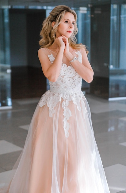 Hübsche junge kaukasische blonde Braut mit Make-up und trendiger Frisur im Hochzeitskleid, die im Innenraum steht und posiert