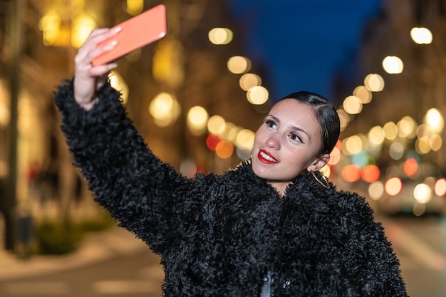 Hübsche junge kaukasische Frau, die nachts mit Licht ein Selfie mit Smartphone macht
