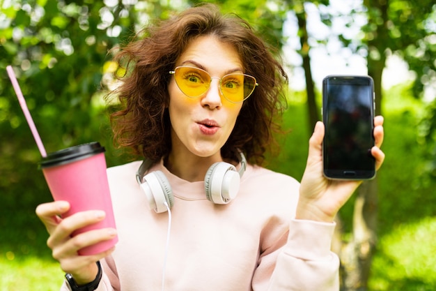 Hübsche junge kaukasische Frau gehen mit einer Tasse Kaffee im Park spazieren und zeigen ihr Telefon