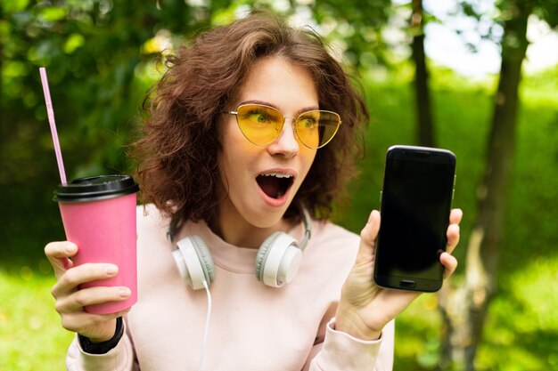 Hübsche junge kaukasische Frau gehen mit einer Tasse Kaffee im Park spazieren und zeigen ihr Telefon