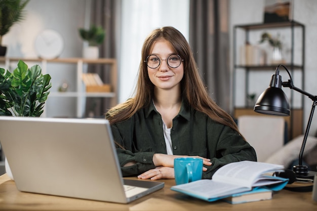 Hübsche junge kaukasische Frau mit langen dunklen Haaren, die mit modernem Laptop am Tisch sitzt