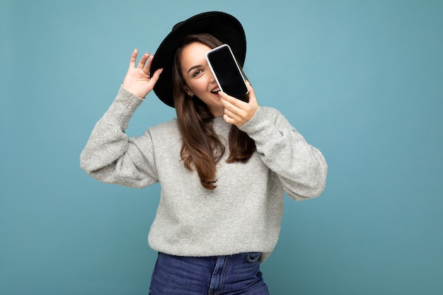 Hübsche junge lächelnde Frau mit schwarzem Hut und grauem Pullover, die das Telefon mit Blick auf die Kamera im Hintergrund hält. Mock-up, Ausschnitt, freier Raum. Platz kopieren