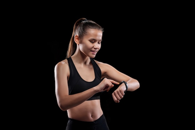 Hübsche junge lächelnde Sportlerin in Aktivkleidung, die auf ihre Smartwatch schaut und auf ihre Anzeige zeigt, während sie isoliert steht
