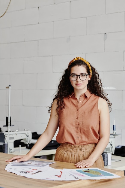 Hübsche junge Modedesignerin, die Skizzen für neue Kollektion wählt, während sie in der Werkstatt gegen Nähmaschinen am Tisch steht