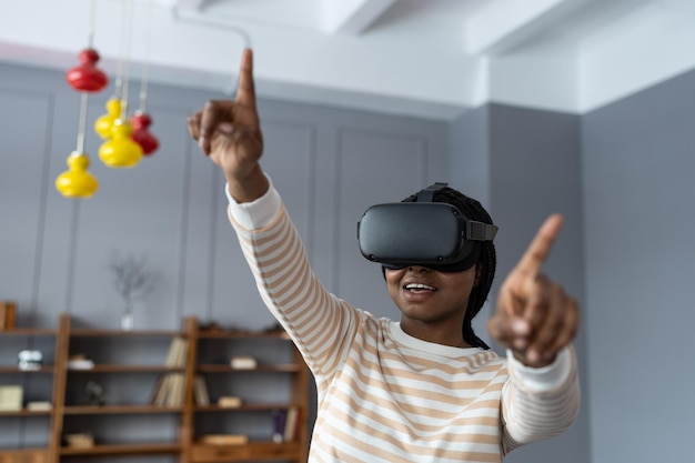 Hübsche junge schwarze Frau im VR-Headset, die Objekte in der Welt der virtuellen Realität anschaut und versucht, sie zu berühren