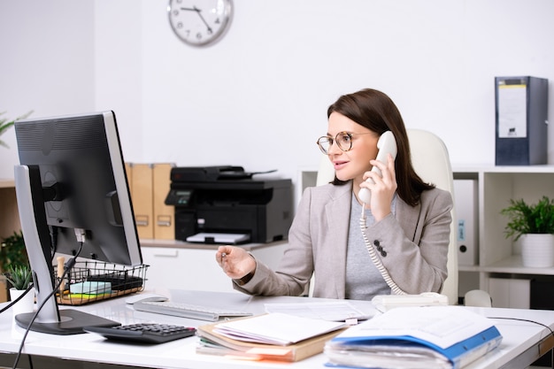 Hübsche junge Sekretärin sitzt am Schreibtisch vor dem Computermonitor, während sie Daten mit dem Kunden am Telefon bespricht
