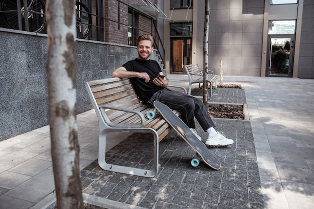 Hübsche junge, stilvolle lächelnde Blicke auf jemanden, während sie das Smartphone benutzen, Beiträge mögen, Inhalte in sozialen Netzwerken erneut teilen, Longboard, das sich auf die Bank lehnt. Typ in lässigem Outfit, Messaging