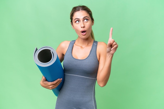 Hübsche junge uruguayische Sportlerin, die zu Yoga-Kursen geht, während sie eine Matte über isoliertem Hintergrund hält und an eine Idee denkt, die mit dem Finger nach oben zeigt