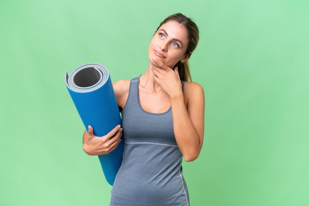 Hübsche junge uruguayische Sportlerin, die zu Yoga-Kursen geht, während sie eine Matte über isoliertem Hintergrund hält und lächelnd nach oben schaut