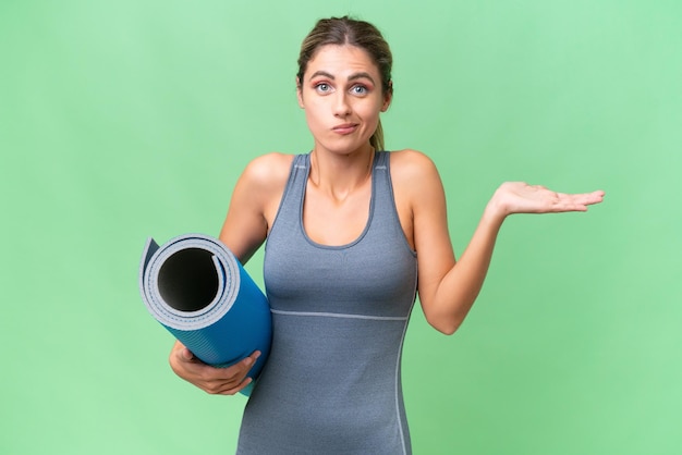 Hübsche junge uruguayische Sportlerin, die zu Yoga-Kursen geht, während sie eine Matte über isoliertem Hintergrund hält und Zweifel hat, während sie die Hände hebt