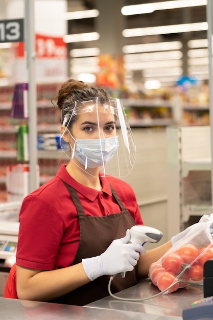 Hübsche junge Verkäuferin in Schürze, die an der Kasse im Supermarkt sitzt und Sie mit einem Lächeln ansieht, während sie Kunden bedient?