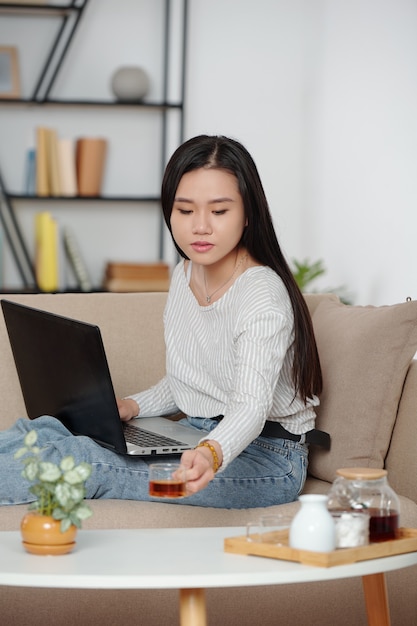 Hübsche junge Vietnamesin, die eine Tasse Tee trinkt und am Laptop arbeitet, wenn sie zu Hause auf dem Sofa sitzt
