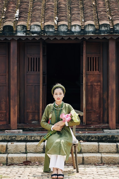 Hübsche junge vietnamesische Frau in traditioneller Kleidung, die auf einem Stuhl auf der Veranda des Tempels mit einem Blumenstrauß aus Lotusblumen sitzt