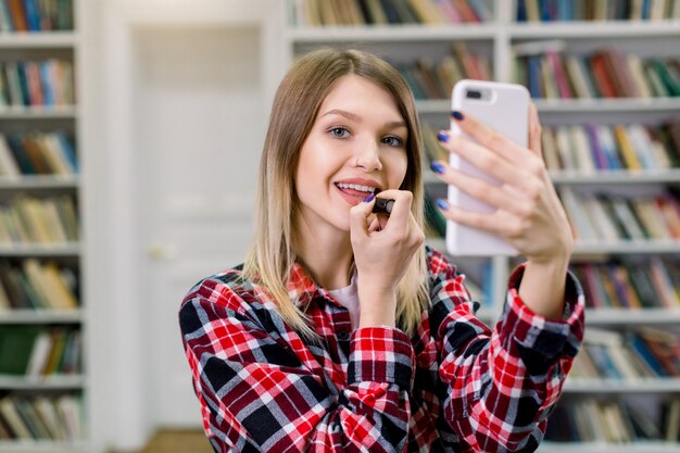 Hübsche kaukasische blonde Studentin im lässigen karierten Hemd, das Make-up in der Bibliothek tut und ihre Lippen mit Lippenstift malt