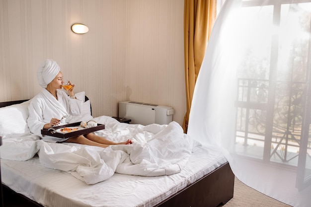 Hübsche kaukasische Frau im Bademantel und Handtuch auf dem Kopf, die morgens im Hotelzimmer im Bett frühstückt