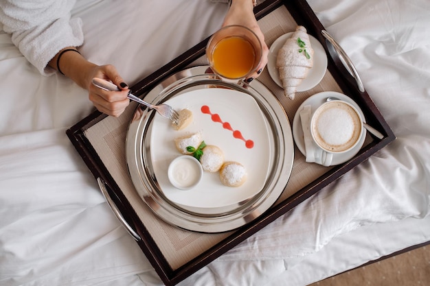 Hübsche kaukasische Frau im Bademantel und Handtuch auf dem Kopf, die morgens im Hotelzimmer im Bett frühstückt