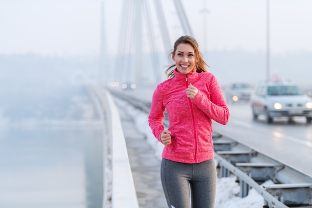 Hübsche kaukasische Frau mit Pferdeschwanz gekleidet in Sportbekleidung, die auf der Brücke im Winter läuft. Gesundes Lebensstilkonzept.