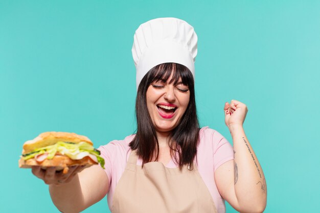 Hübsche Kochfrau, die erfolgreich einen Sieg feiert und ein Sandwich hält