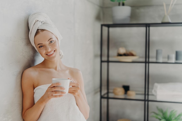 Hübsche lächelnde Frau mit gesunder Haut, die in ein weißes Handtuch gehüllt ist, steht froh, dass Kaffee im Innenbereich einen positiven Gesichtsausdruck hat und Posen im Innenbereich darstellt. People Spa verwöhnt Hautpflege- und Wellnesskonzept