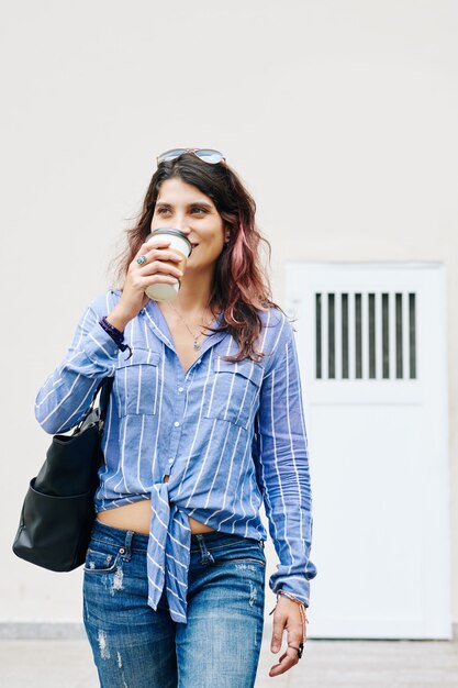 Hübsche lächelnde junge Frau im gestreiften Hemd, die Kaffee trinkt und die Straße entlang geht