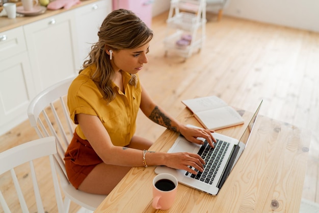 Hübsche lächelnde Studentin, die Laptop benutzt und Notizen über ihre moderne helle Küche schreibt