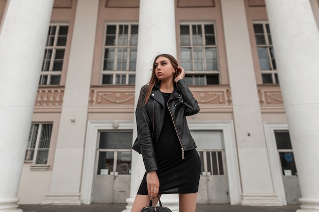 Hübsche modische junge Frau in einer trendigen schwarzen Lederjacke in einem eleganten Kleid mit einer stilvollen Tasche steht in der Nähe einer weißen Vintage-Säule auf der Straße. Modernes europäisches Mädchen. Lässige Herbstmode.