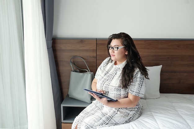 Hübsche nachdenkliche junge Frau in Übergröße, die mit Tablet-Computer auf dem Bett sitzt und durch das Fenster schaut