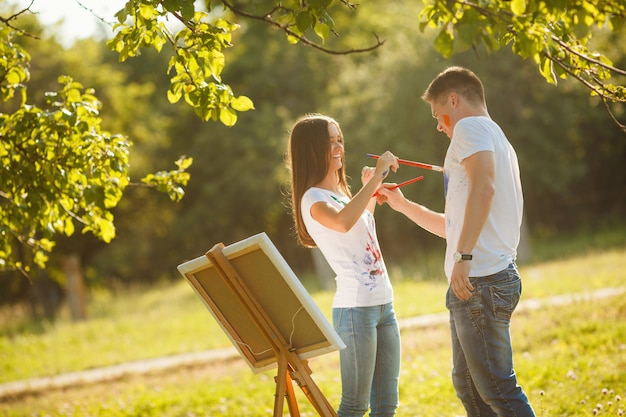 Hübsche Paare, die Spaß am Freilicht draußen haben. Junger Mann und Frau, die durch bunte Farben auf einander `s T-Shirts mit Bürsten nahe dem Gestell auf der Natur zeichnet.