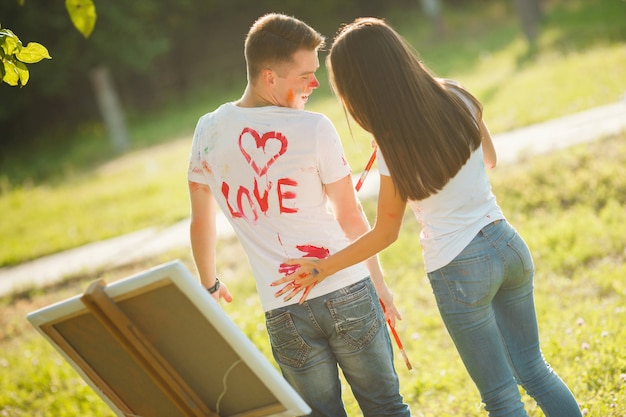 Hübsche Paare, die Spaß am Freilicht draußen haben. Junger Mann und Frau, die durch bunte Farben auf einander `s T-Shirts mit ihren Händen nahe dem Gestell auf der Natur zeichnet.