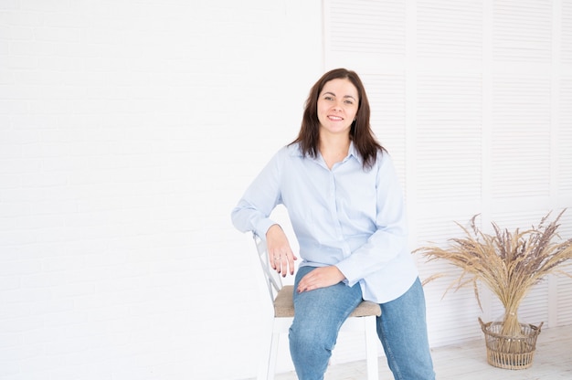 Hübsche Plus-Size-Model-Frau in Hemd und Jeans posiert im Studio auf Stuhl, weißer Hintergrund mit Kopienraum.